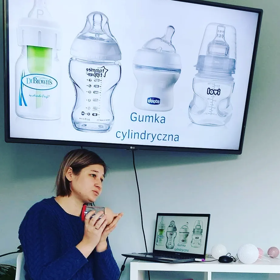 Kobieta prowadzi szkolenie, siedząc przy biurku z laptopem pod dużym monitorem. Na ekranie głównym wyświetlony jest napis "Gumka cylindryczna" oraz zdjęcia czterech różnych butelek do karmienia niemowląt marek Dr Brown's, Tommee Tippee, Chicco i Lovi. Kobieta trzyma w dłoniach model anatomiczny do demonstracji.