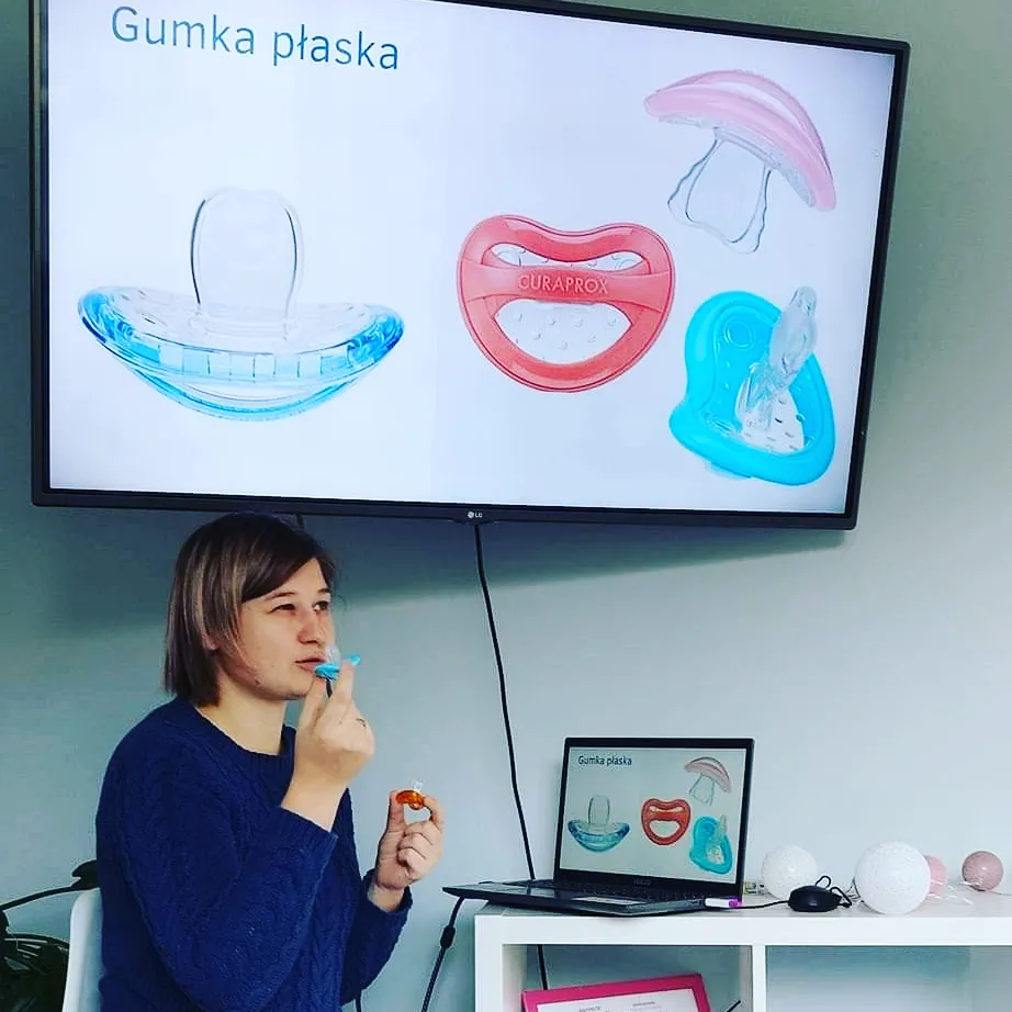 Kobieta siedzi w pokoju przed laptopem i dużym ekranem telewizora, prowadząc prezentację. Na ekranie widnieje napis "Gumka płaska" oraz ilustracje różnych rodzajów smoczków uspokajających. Kobieta trzyma w dłoniach małe akcesoria do karmienia, demonstrując je.