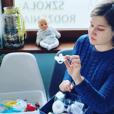 Kobieta w niebieskim swetrze siedzi przy stole i trzyma przezroczysty smoczek, przyglądając mu się uważnie. Na parapecie za nią siedzi lalka niemowlęca, a obok stoją pojemniki z różnymi akcesoriami logopedycznymi i smoczkami.
