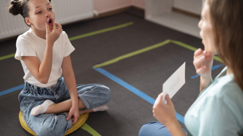 Młoda dziewczynka i dorosła kobieta siedzą naprzeciwko siebie na podłodze w gabinecie terapeutycznym. Obie wykonują ćwiczenia logopedyczne, dotykając palcem ust i układając je w kształt litery "o". Kobieta trzyma w dłoni białą kartkę, prawdopodobnie z instrukcją lub obrazkiem.